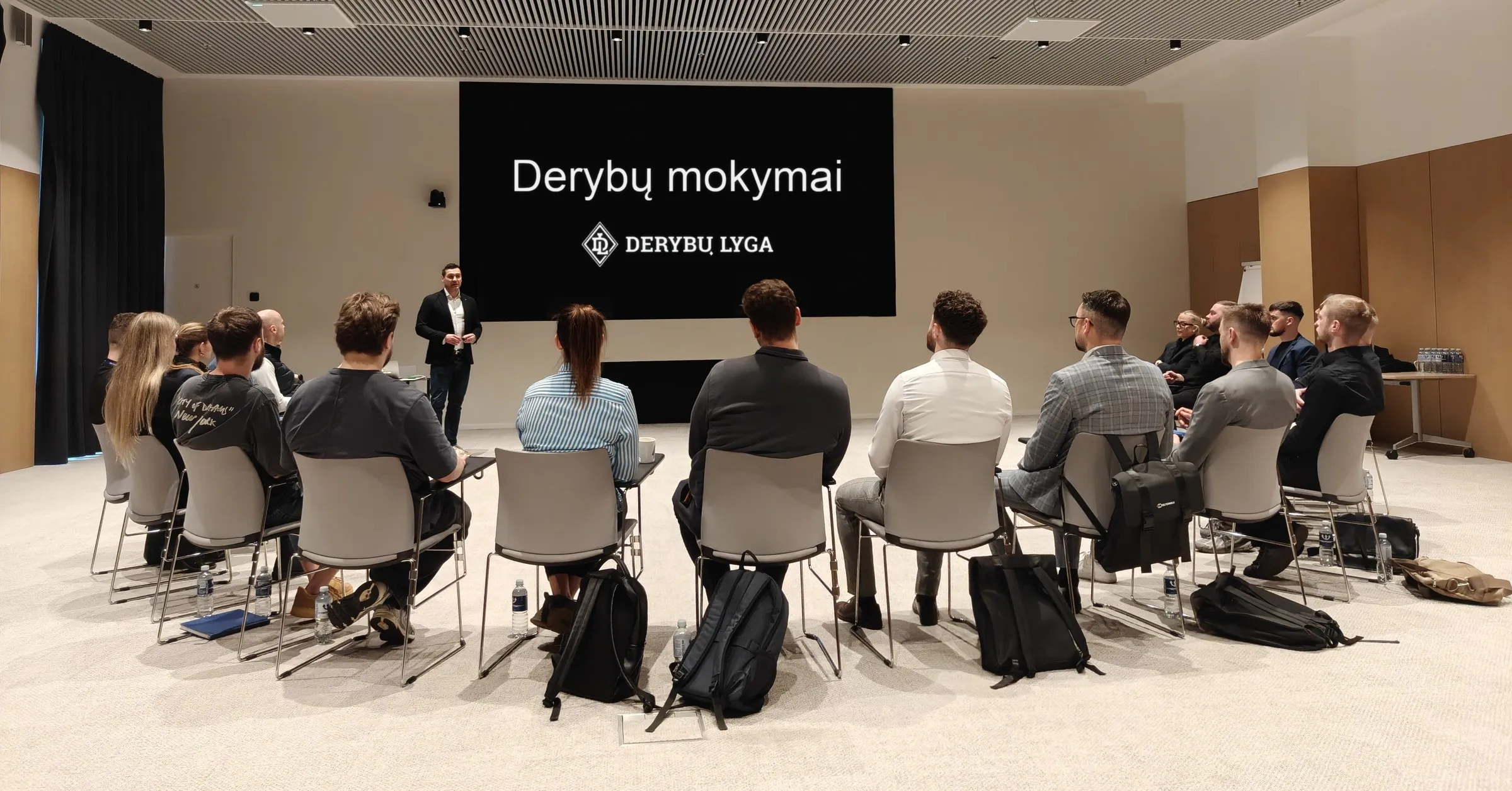 Derybų mokymų dalyviai ratu klausosi trenerio prie ekrano su užrašu „Derybų mokymai“